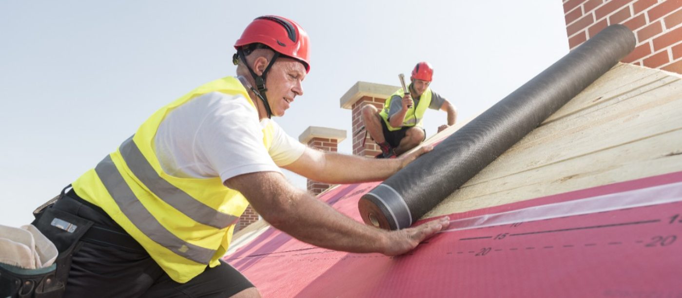 Urban roofers applying roof underlay sheet wearing hard hat and safety jacket brick chimney